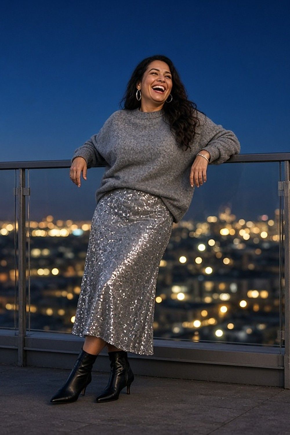 Full-length photo of a laughing woman leaning on a rooftop railing at dusk above city lights, in an oversized grey cashmere jumper, a silver sequin midi skirt and black pointed kitten-heel ankle boots.
